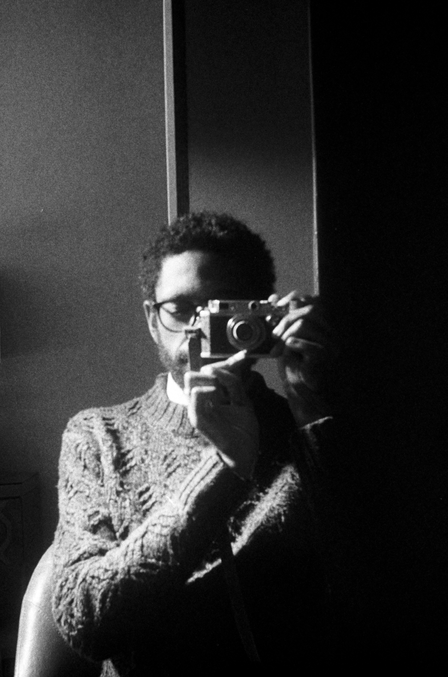 Sean Page Collins in a black and white photo. Sean has a short Afro, black glasses, and a beard. The left half of his face is in shadow, and is covered by a Cannon IID camera. He wears a crew neck sweater with a diamond pattern. He seems to have taken a photo of himself in the mirror. He is seated; the wall behind him is dark.