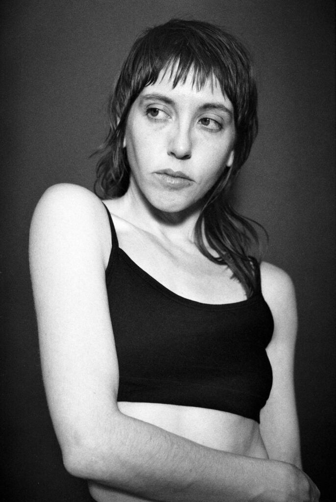 A black and white photo: A slim white woman with a shaggy haircut purses her lips, making the lines of her cheekbones pop. She wears a black spaghetti strap tank and is in front of a grey background.