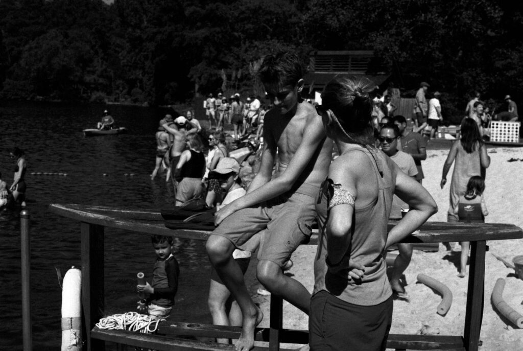 A black and white photo: A slice of light illuminates the body of a white boy, perhaps 13, in swim trunks. He sits on a railing, his head bowed, largely in shadow. A woman, her hands on her hips, faces him and speaks to him.