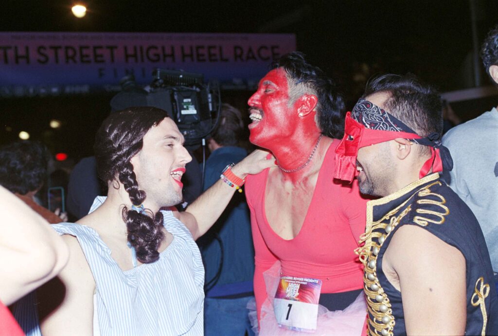 At night, lit by flash, a man, his face and neck painted red, sweat making him shiny, tilts his head back, mouth open. A man in a blue dress, wearing brown pigtails and a red lip places his hand on the red-faced man's shoulder. A third man, in a navy vest with yellow ribbons down the from and a red mask smiles at them both.