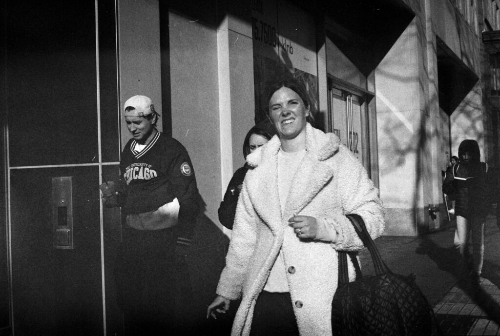 In a black and white photo: A white woman, dark hair pulled back, squints at the camera, one eye open, one closed, her fuzzy white coat shining in the sun. Behind her, a white teen boy half wears a University of Chicago jersey, its bottom rolled up to reveal the white t-shirt he has on underneath.